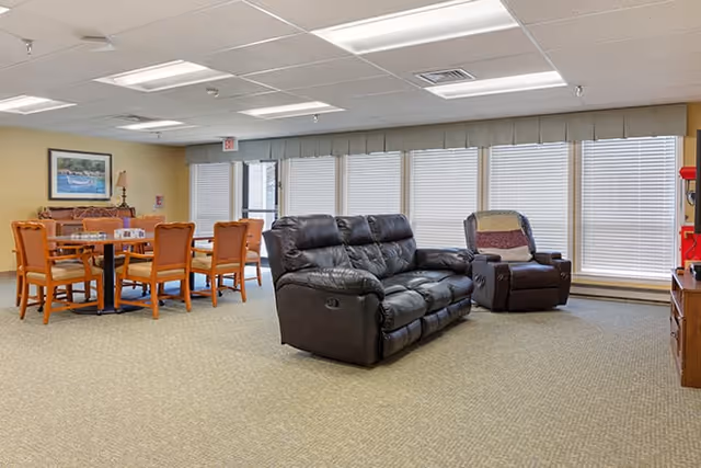 A spacious senior living common area with a black leather reclining sofa and matching armchair. There are several wooden tables and chairs in the background near large windows with blinds. The room has beige carpet and light-colored walls with ceiling lights.