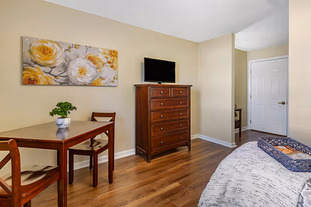 Furnished bedroom with a wooden dresser and TV, a small dining table with chairs, floral wall art, and hardwood floors.