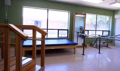Rehabilitation room with a padded therapy mat, parallel bars, and large windows.