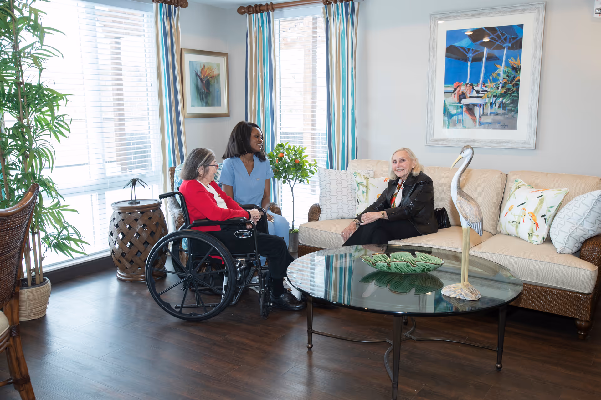 A bright living room with large windows and striped curtains. An elderly woman in a wheelchair, a caregiver in blue scrubs, and another elderly woman sitting on a beige couch are engaged in conversation. The room features a glass coffee table with a decorative bird sculpture and a green dish, a potted plant, and framed artwork on the walls.