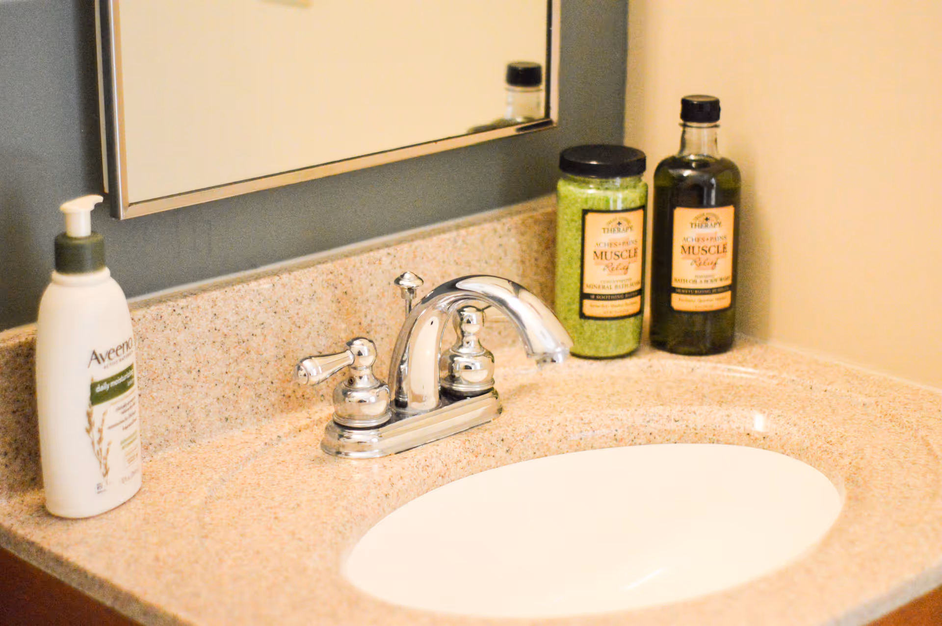 A bathroom sink with a chrome faucet on a beige countertop. To the left of the faucet is a bottle of Aveeno daily moisturizing lotion, and to the right are two bottles of muscle relief bath products. A mirror is mounted on the wall above the sink.