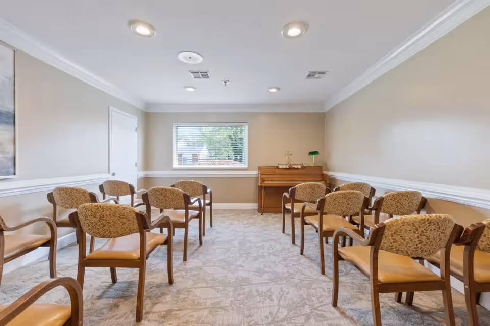 A small meeting or activity room with beige walls and carpeted floor. There are two rows of wooden chairs with cushioned seats and backs arranged facing a wooden piano against the far wall. A window with blinds is centered on the back wall, letting in natural light. The ceiling has recessed lighting and ventilation fixtures.