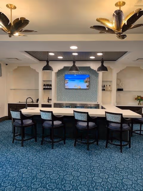 Interior view of a modern kitchen area with a large white island countertop surrounded by five cushioned bar stools. Above the island, there are three black pendant lights. The back wall features a mounted flat-screen TV displaying a beach scene, with shelves on either side holding bottles and decorative items. The ceiling has two decorative gold palm leaf light fixtures, and the floor is covered with a blue patterned carpet.