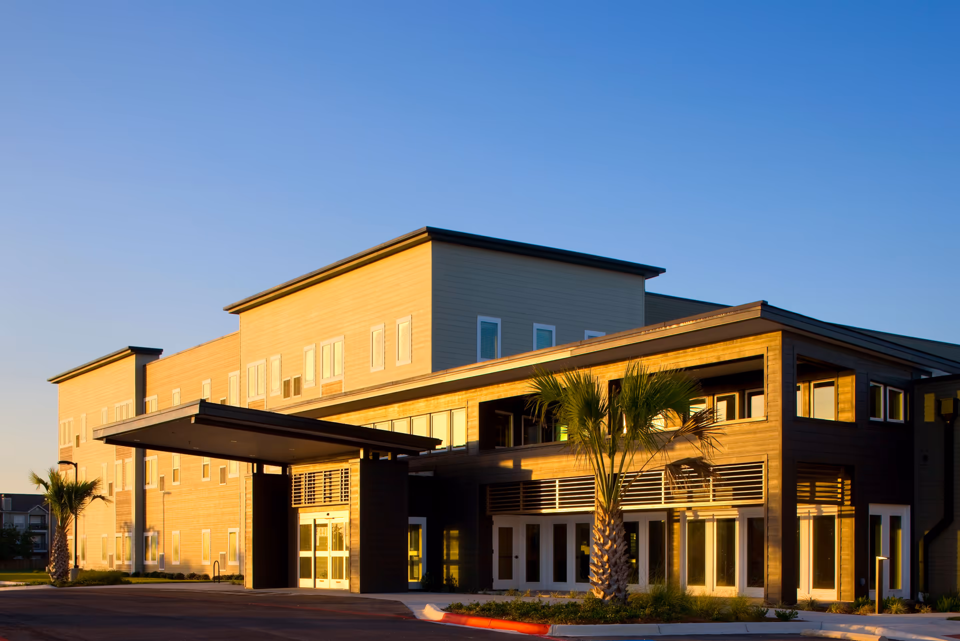 Exterior view of Elan Corpus Christi senior living facility with a two-story tan and brown facade and a covered entrance.