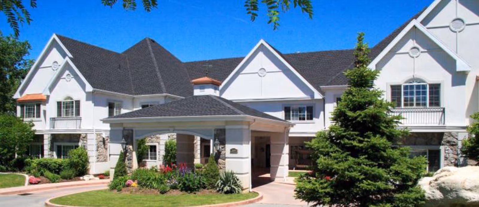 Exterior view of a senior living facility building with white walls, dark shingled roof, and a covered entrance. The surrounding area is landscaped with green grass, bushes, flowers, and a large evergreen tree under a clear blue sky.