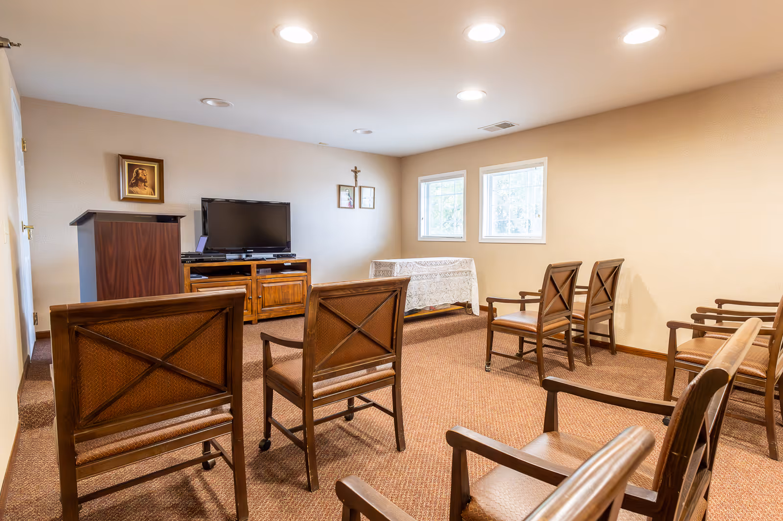 A small room with several wooden chairs arranged in rows facing a TV on a wooden stand. There is a wooden podium to the left and a table covered with a white lace cloth near two windows on the right wall. The walls are beige and the floor is carpeted in brown.