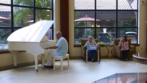 A man playing a white grand piano in a spacious room with large windows. Two women are seated nearby, one in a wheelchair and the other on a chair, both watching the pianist. Outside the windows, there are umbrellas and greenery visible.