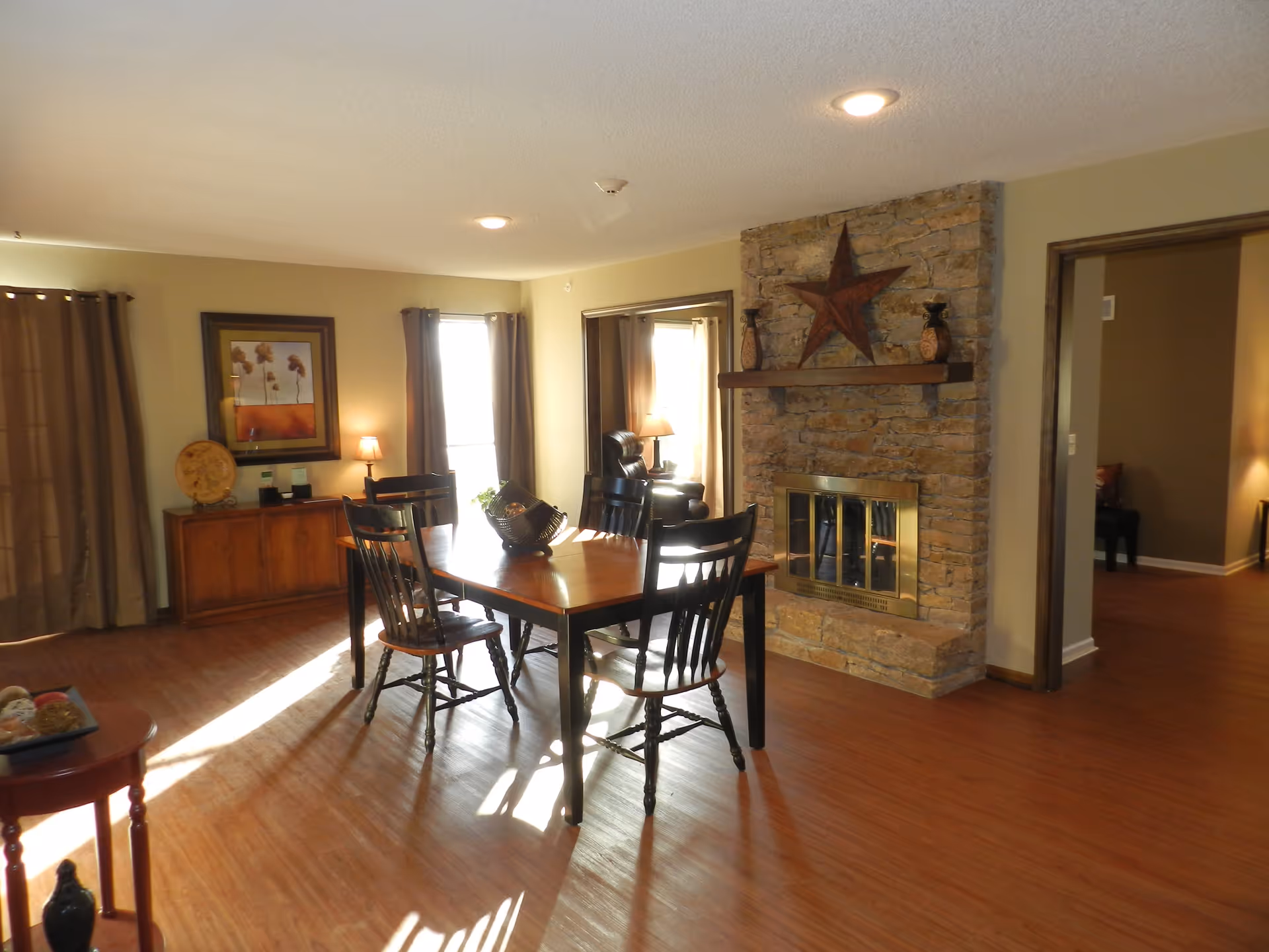 A cozy dining area with a wooden table and four black chairs, a stone fireplace with a large star decoration above it, and a sideboard with a framed picture and lamp. The room has wooden flooring and beige walls with windows covered by brown curtains allowing sunlight to stream in.