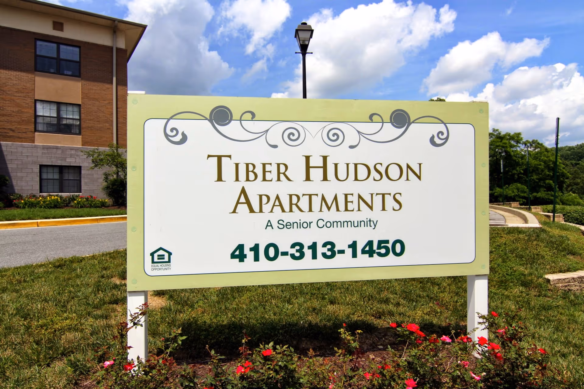 Outdoor view of a sign for Tiber Hudson Apartments, a senior community, with a phone number 410-313-1450. The sign is placed on a grassy area with flowers in front and a building partially visible in the background under a partly cloudy sky.