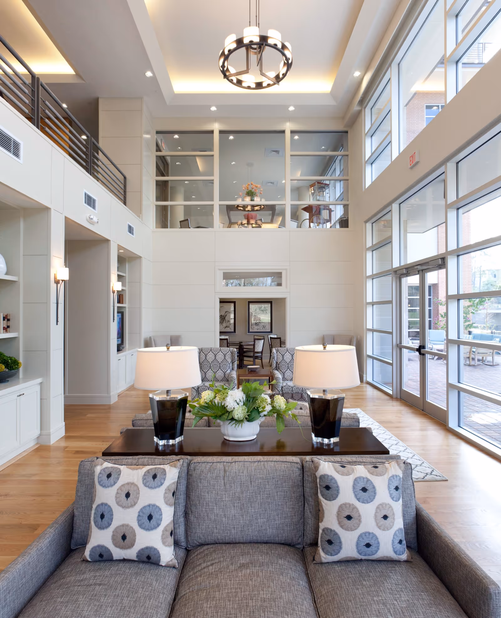A bright and spacious living room area with large floor-to-ceiling windows on the right side allowing natural light to fill the space. The room features a gray sofa with two patterned pillows, a dark wooden table behind it with two black lamps and a white flower arrangement. In the background, there are two patterned armchairs and a dining area with chairs and framed artwork on the wall. The ceiling has recessed lighting and a modern chandelier, with a second-floor balcony visible above.