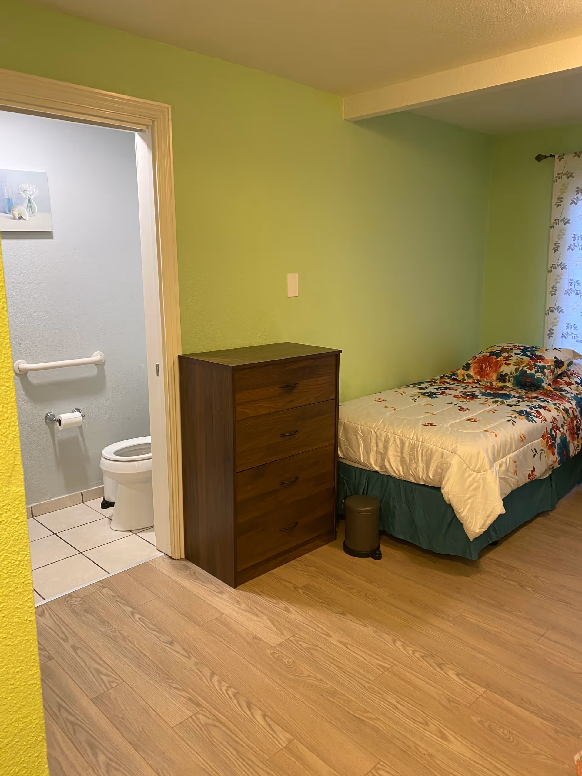 A small bedroom with light green walls and wood flooring. There is a single bed with a floral bedspread and a matching pillow. Next to the bed is a dark wooden chest of drawers and a small trash can. An open door reveals a bathroom with a toilet, grab bar, and toilet paper holder, with light blue walls and tiled floor.