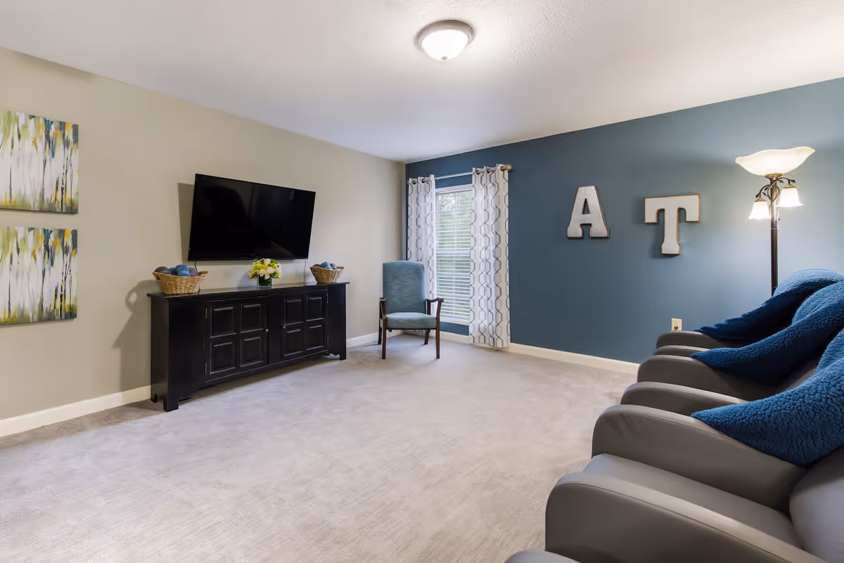 A living room with a beige carpet and walls painted beige and blue. There is a black TV stand with a flat-screen TV mounted above it, two baskets, and a flower arrangement. A single blue armchair is placed near a window with patterned curtains. On the blue wall, large decorative letters 'A' and 'T' are mounted. A floor lamp with multiple lights stands next to a row of gray recliner chairs with blue cushions.