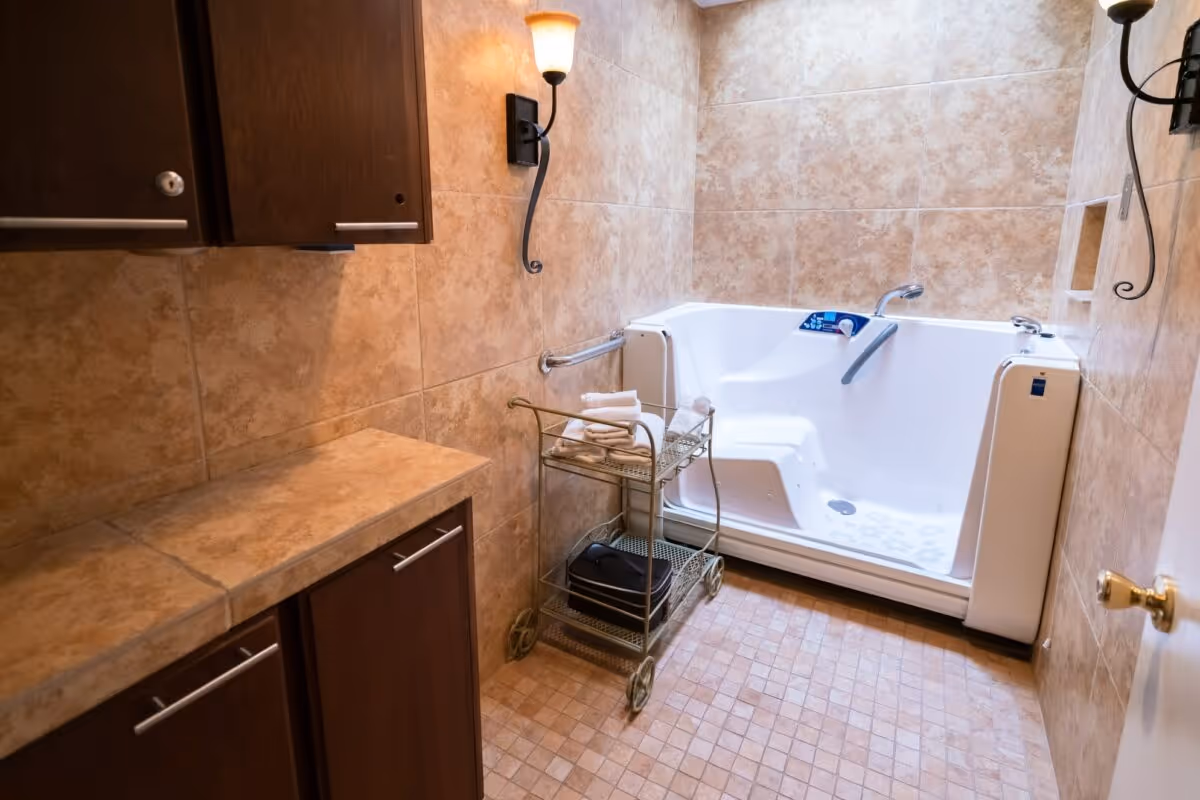 A bathroom with beige tiled walls and floor featuring a white walk-in bathtub with safety grab bars. There is a metal cart with towels and other items next to the tub, and dark wooden cabinets with silver handles on the left wall. Two wall-mounted light fixtures provide warm lighting.
