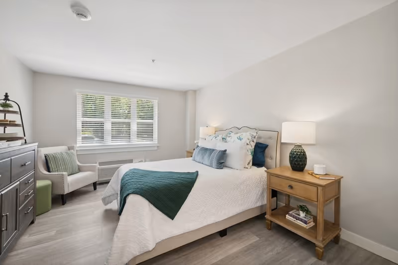 A bright and cozy bedroom with a large window letting in natural light. The room features a neatly made bed with white bedding and teal accents, a wooden nightstand with a lamp and books, a cushioned armchair with a striped pillow, and a dark wooden dresser with decorative items.