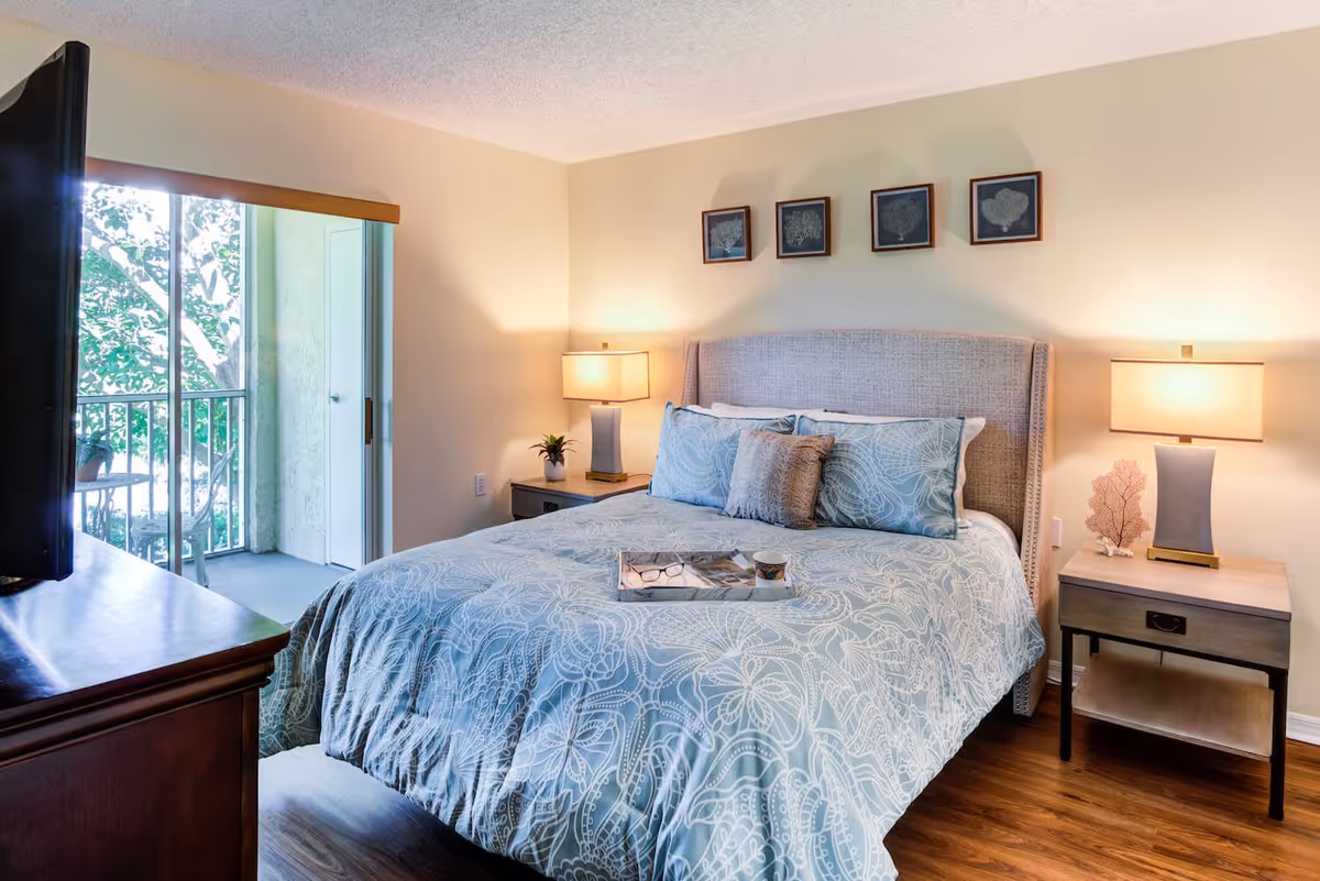 Bright furnished bedroom with a bed dressed in patterned bedding, bedside tables and lamps, a TV on a dresser, and a sliding glass door to a small balcony.