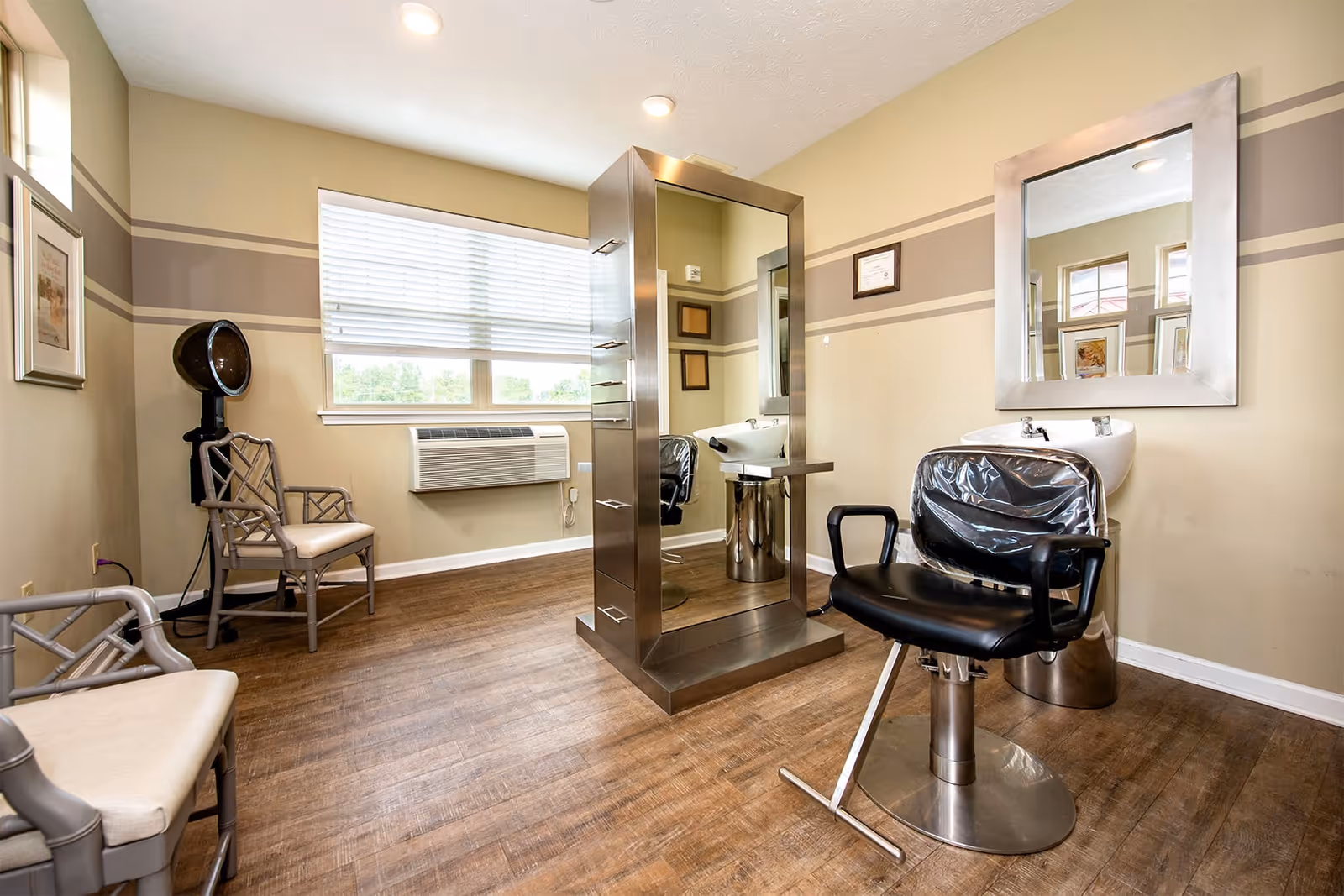 Interior of a hair salon area in a senior living facility with a black salon chair in front of a wash basin and a large mirror. There is a central mirrored storage unit, two beige chairs, a wall-mounted air conditioning unit, and framed pictures on the beige walls with horizontal stripes.