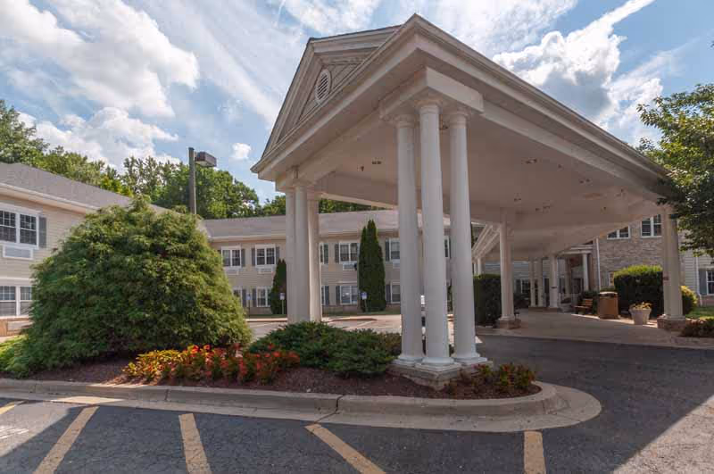 Entrance area of a senior living facility with a covered driveway supported by white columns, surrounded by landscaped bushes and flowers, with a two-story building in the background under a partly cloudy sky.