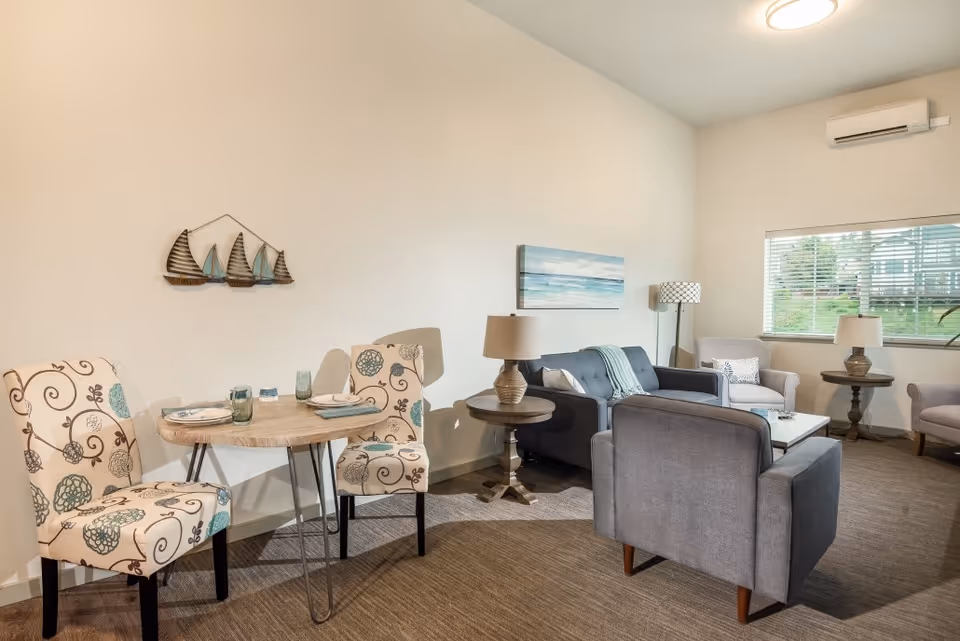 A cozy living room area with a small round dining table set for two with patterned chairs, a gray sofa, two gray armchairs, a coffee table, side tables with lamps, a floor lamp, a wall-mounted air conditioner, a window with blinds, and wall decorations including a painting of the ocean and a metal sailboat sculpture.