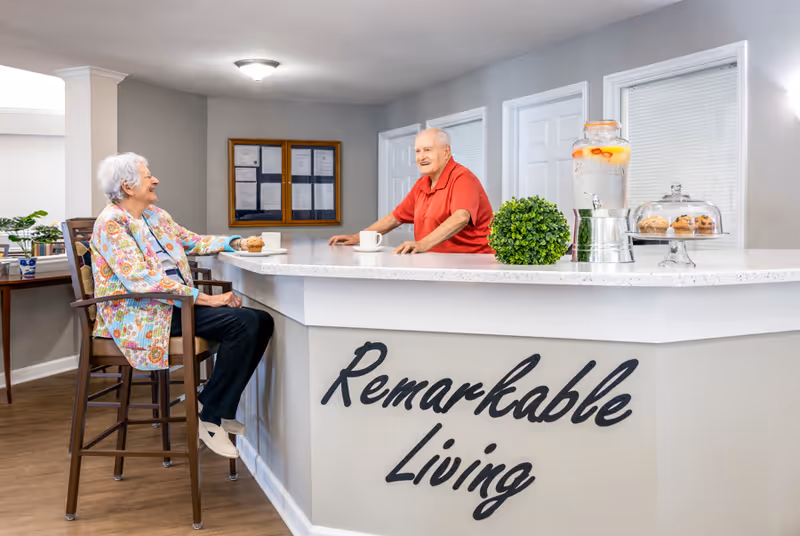 Two elderly residents chat across a reception-style counter in a senior living facility with a 'Remarkable Living' sign.