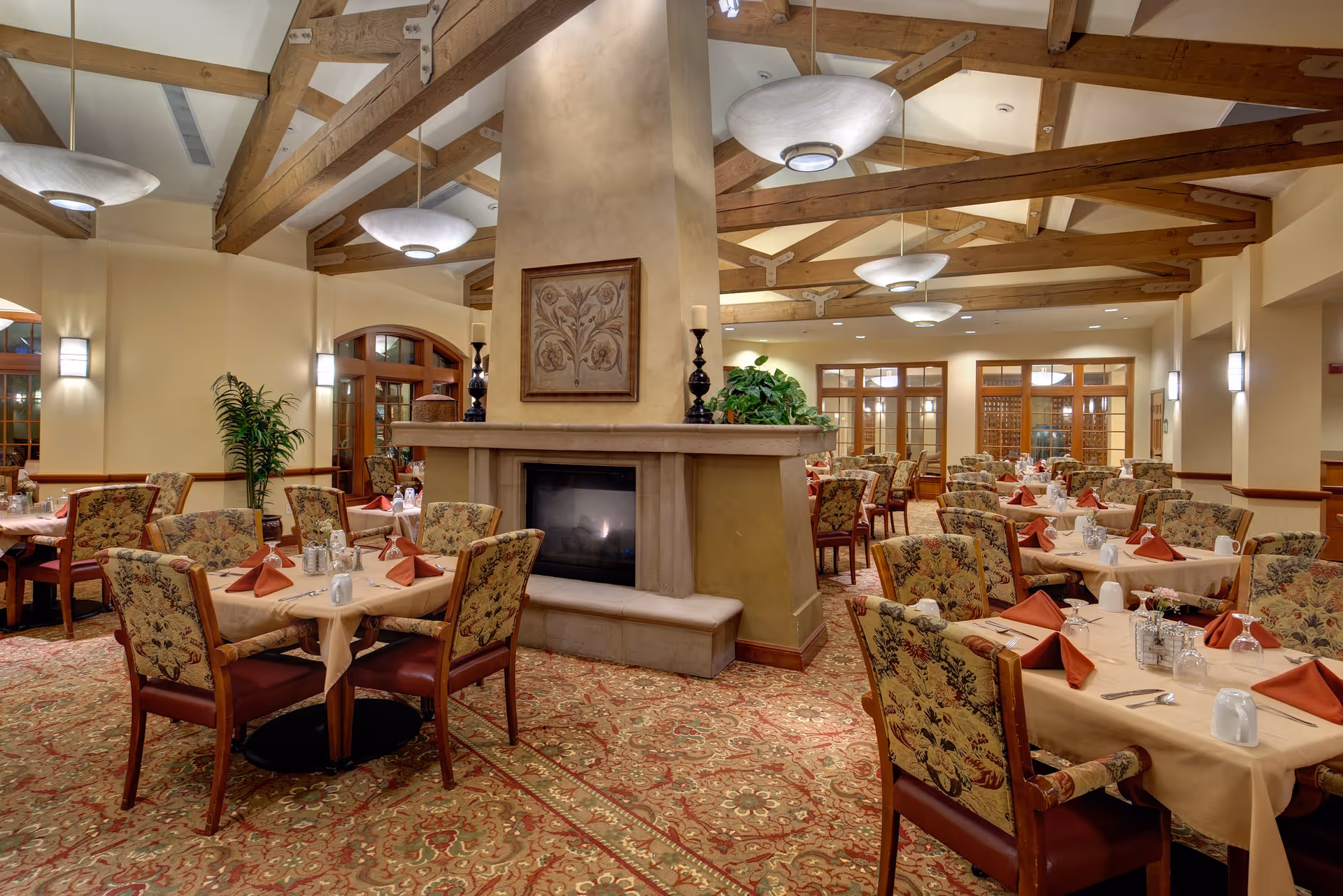 A spacious dining room with multiple tables covered in beige tablecloths, each set with red folded napkins, glasses, and silverware. The room features a large central fireplace with decorative artwork above it, wooden ceiling beams, elegant light fixtures, and patterned carpet. There are upholstered chairs with floral patterns around the tables and some green plants near the walls.