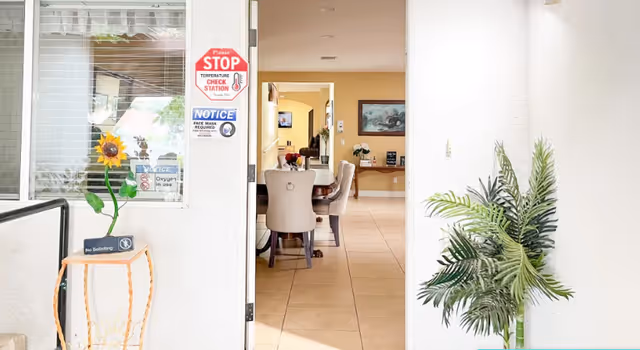 View through an open doorway into a bright interior room with a dining table and chairs. A potted plant is on the right side of the doorway, and a small table with a sunflower decoration and a 'No Soliciting' sign is on the left. Several signs are posted on the wall near the door, including a red stop sign indicating a temperature check station and a notice about face mask requirements.