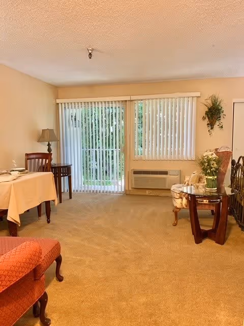 A cozy living room area with beige carpet and walls. There is a sliding glass door with vertical blinds leading outside, and a window with matching blinds. The room features a small dining table with two chairs, a floor lamp, an upholstered armchair, and a side table with a flower arrangement. An air conditioning unit is installed below the window.