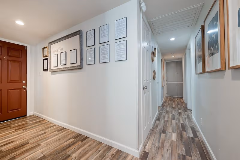 A clean interior entry hallway with a red front door, framed wall art, and wood-look flooring leading to a long corridor.