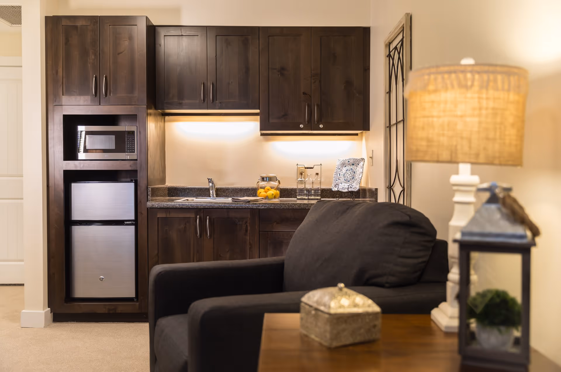 A cozy interior space featuring a small kitchenette with dark wooden cabinets, a microwave, and a mini refrigerator. In the foreground, there is a black cushioned armchair next to a wooden side table with a decorative box, a white lamp with a woven lampshade, and a small lantern with a plant inside.