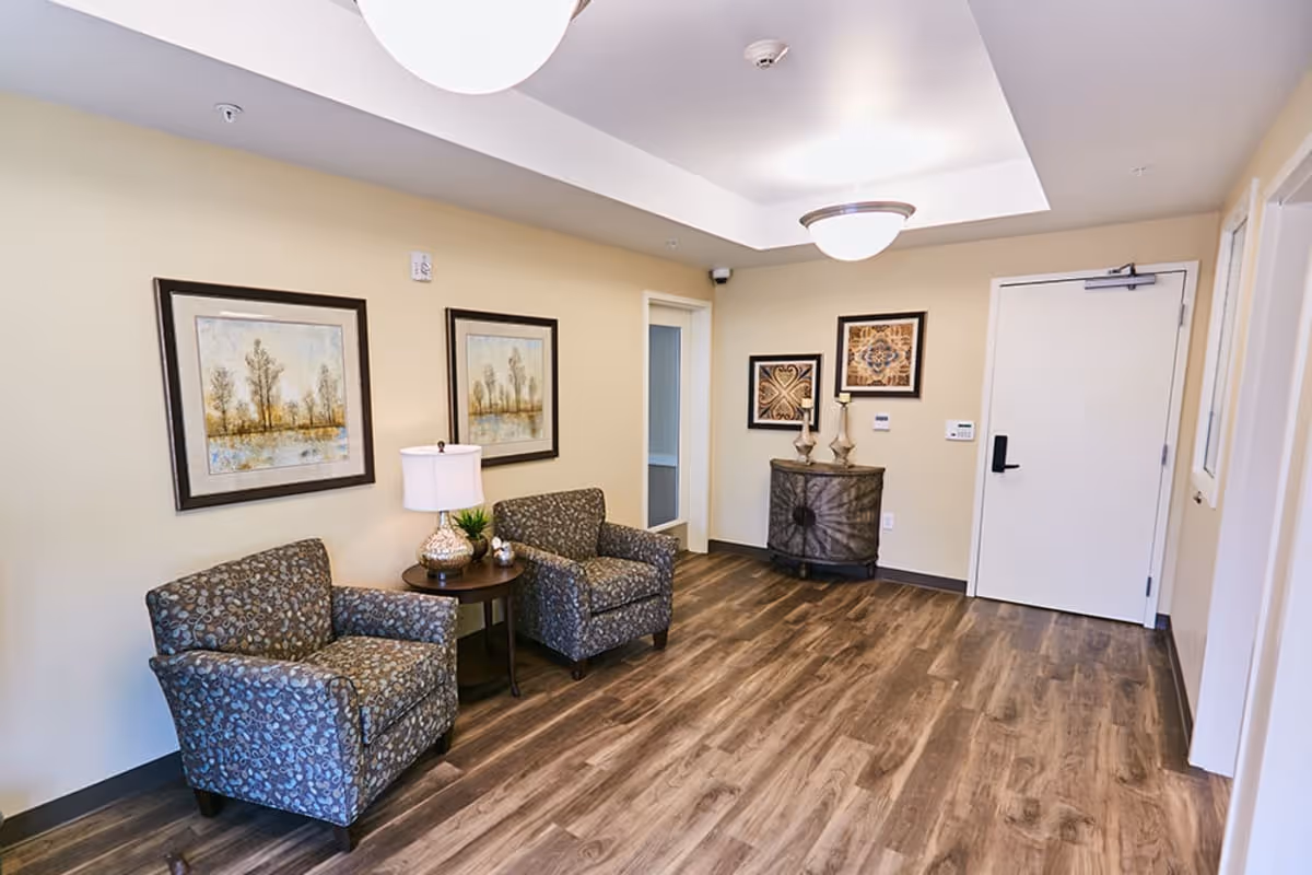 Small lobby seating area with two patterned armchairs, a side table and lamp, framed artwork, and a white entry door.