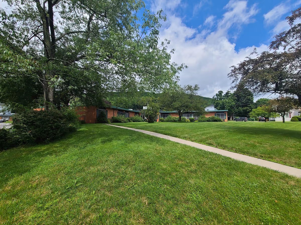 Brick single-story nursing center building set behind a wide grassy lawn with trees and a concrete walkway under a partly cloudy sky.