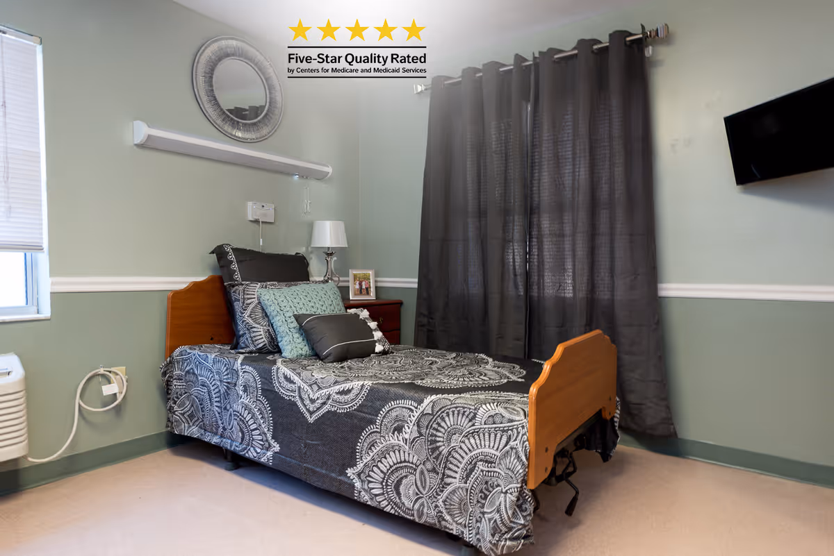 A small, neatly arranged bedroom in a rehabilitation and nursing center featuring a single bed with a patterned black and white bedspread and multiple pillows. The room has light green walls with a white chair rail, a window with closed blinds on the left, dark curtains covering another window, a wall-mounted TV on the right, a bedside table with a lamp and a small framed photo, and a round decorative mirror above the bed. A five-star quality rating graphic is displayed on the wall above the bed.