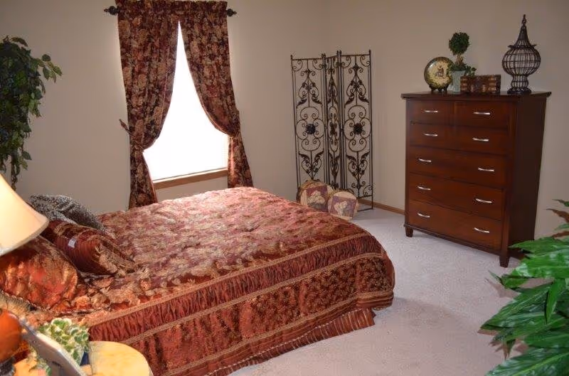 A bedroom with a large bed covered in a red and gold patterned comforter and matching pillows. There is a window with red patterned curtains, a wooden dresser with decorative items on top, a decorative metal room divider, and some green plants in the corners of the room.