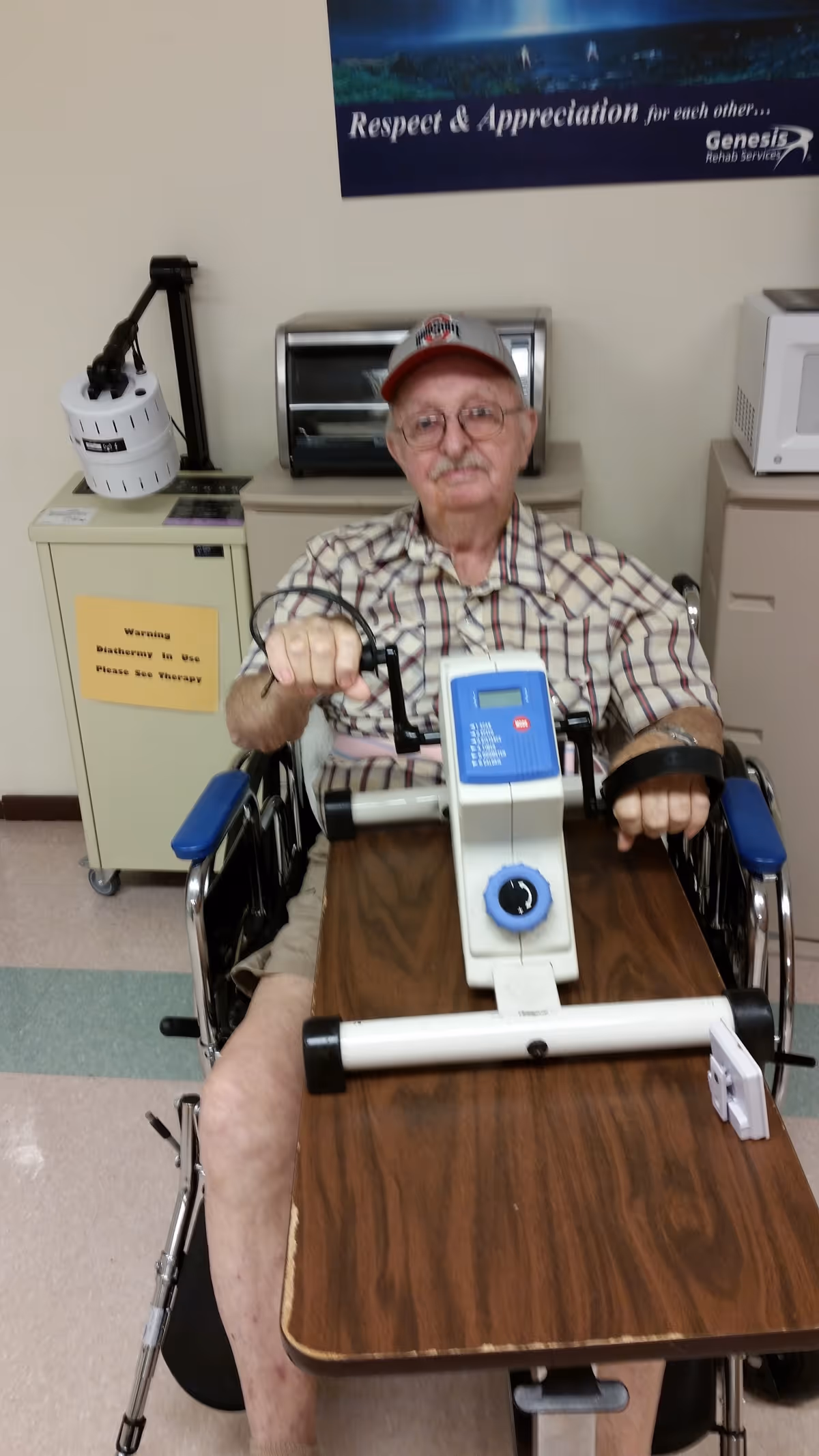 An elderly man wearing a plaid shirt and a baseball cap is seated in a wheelchair using a hand and arm exercise machine placed on a wooden table in front of him. Behind him, there is a cabinet with a toaster oven and a microwave on top. A poster on the wall reads 'Respect & Appreciation for each other... Genesis Rehab Services.'