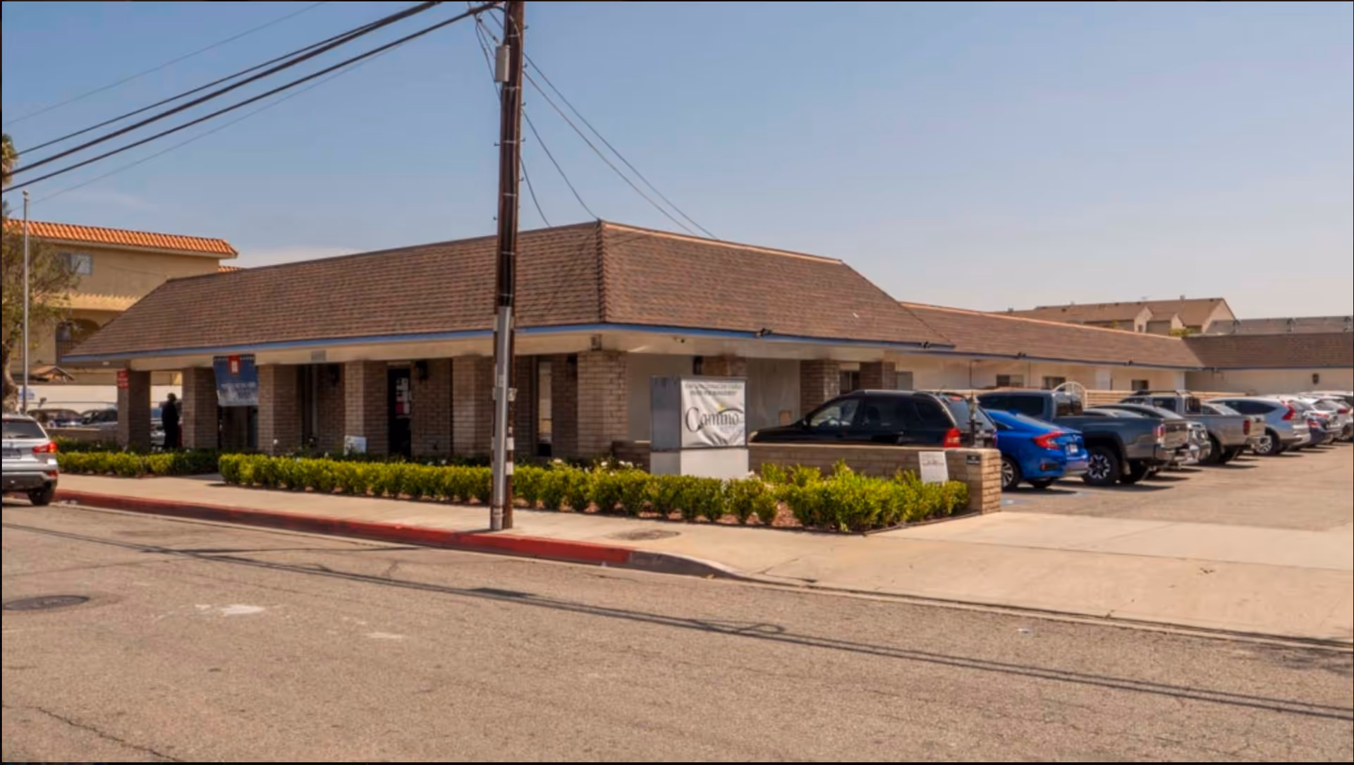 Exterior view of Camino Healthcare facility showing a single-story brick building with a brown shingle roof, a parking lot filled with cars, and a clear blue sky.