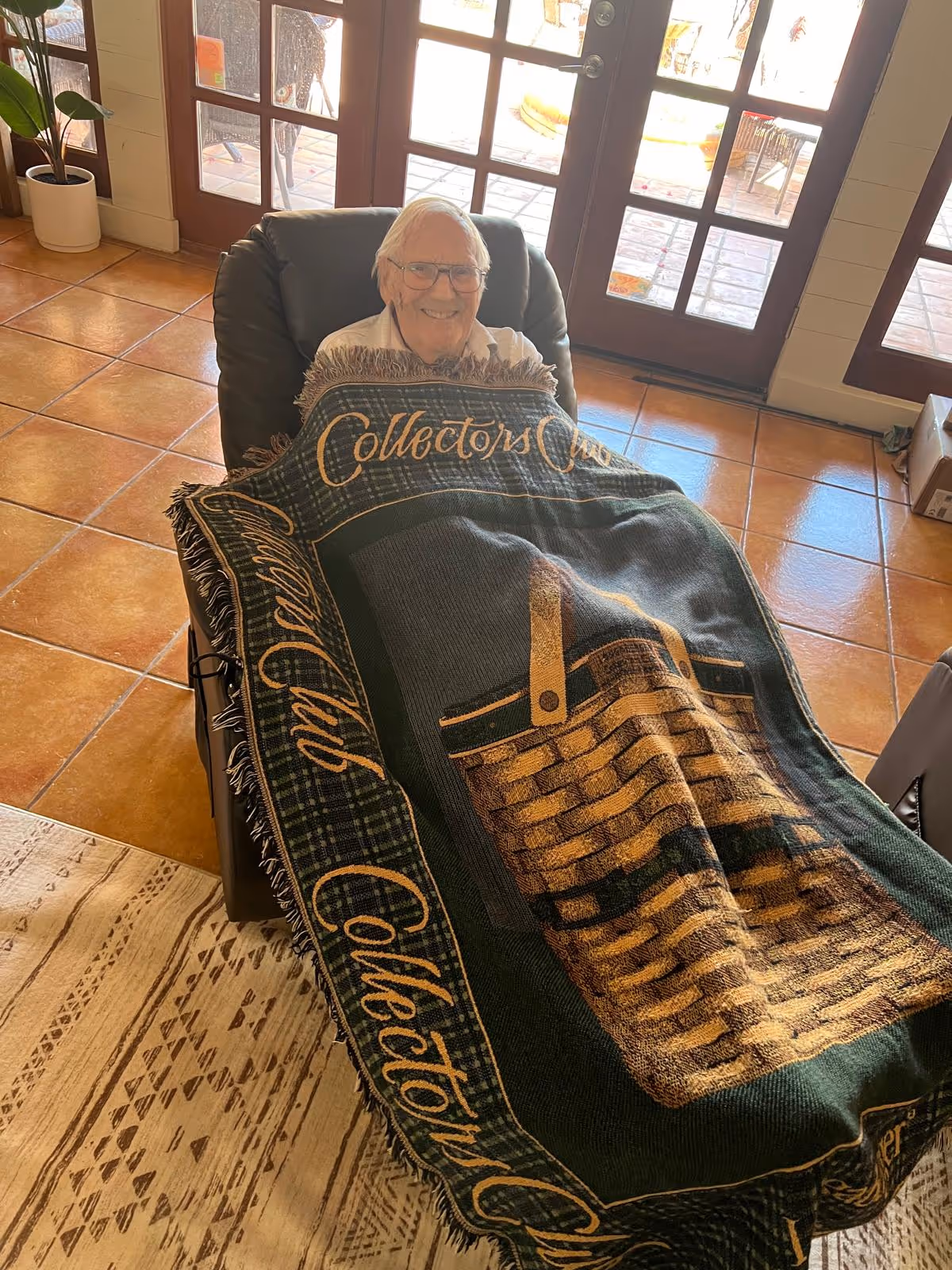 An elderly man smiles while seated in a recliner covered by a 'Collectors Club' blanket in a sunlit living room near French doors.
