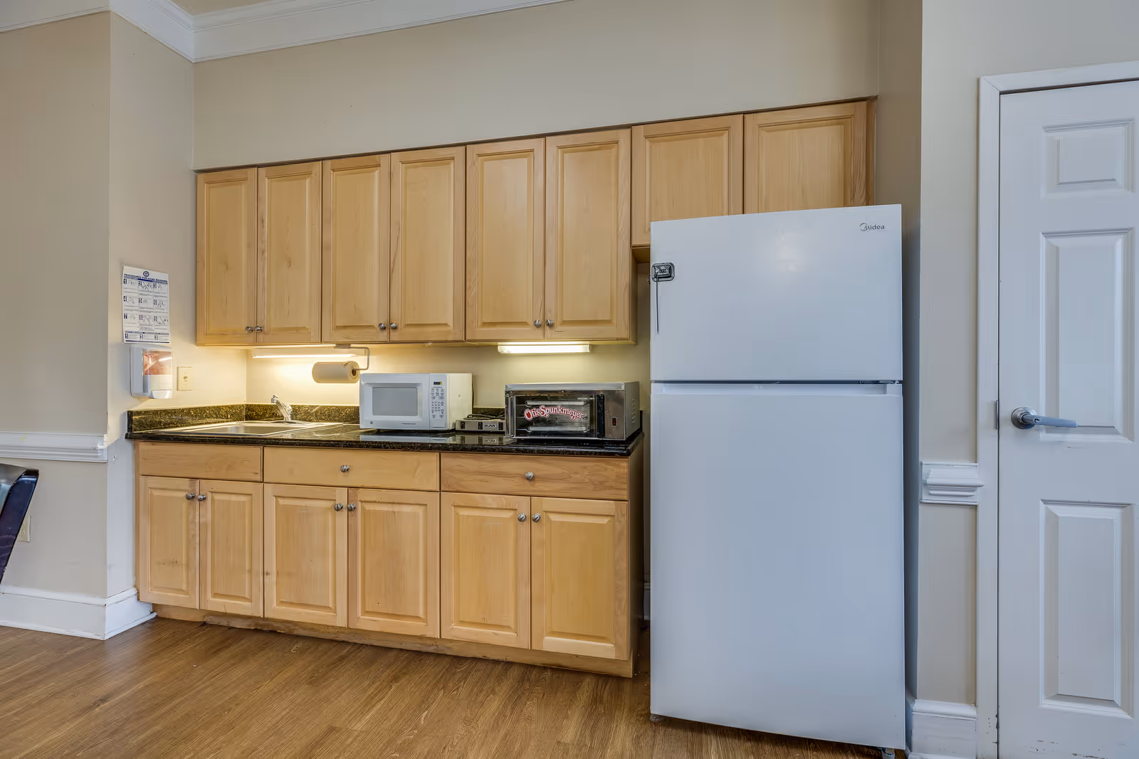 A small kitchen area with light wood cabinets, a black countertop, a white microwave, a toaster oven, and a white refrigerator. The floor is wooden, and there is a white door to the right of the refrigerator. Under-cabinet lighting illuminates the countertop.