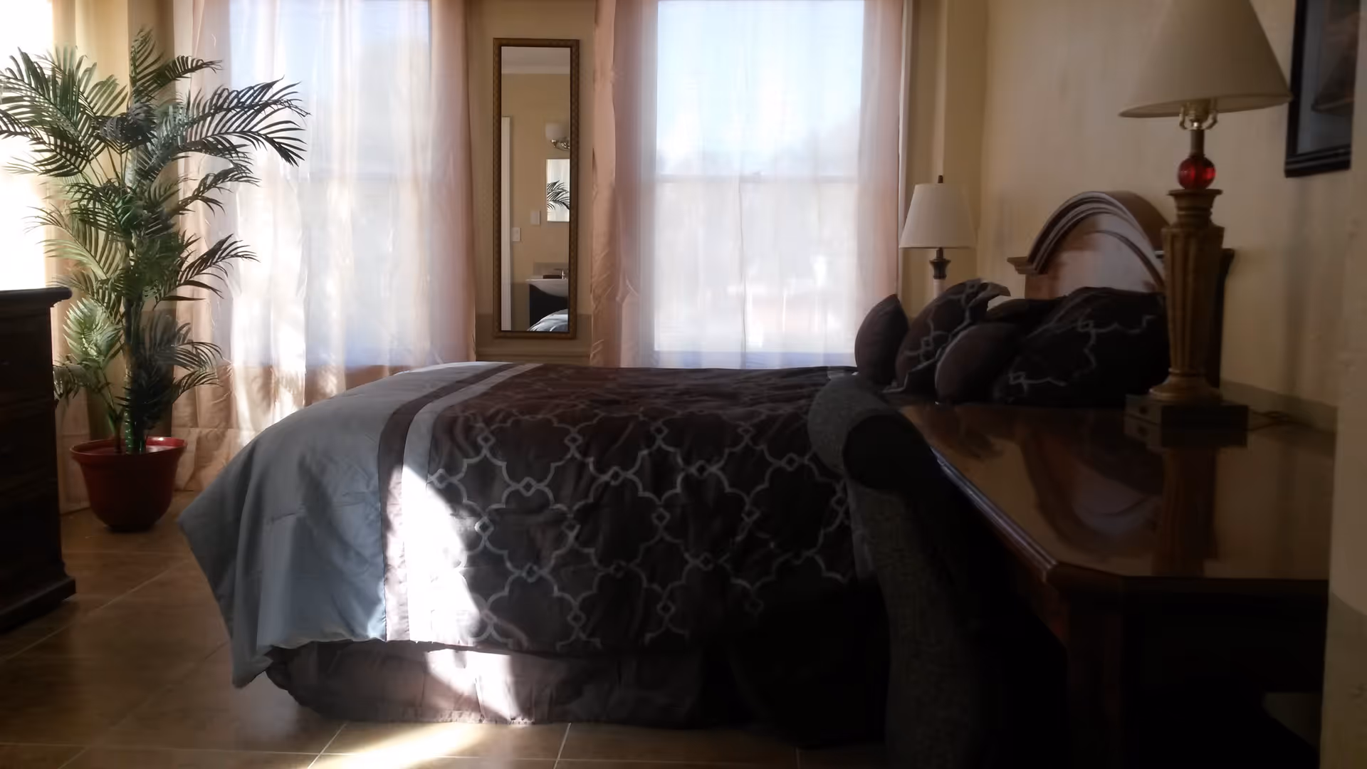 A bedroom with a bed covered in a dark patterned comforter and several pillows. There is a wooden headboard, a wooden desk with a lamp on it, and a chair. Two large windows with sheer curtains allow natural light to fill the room. A tall potted plant is placed near the window, and a full-length mirror hangs on the wall between the windows.