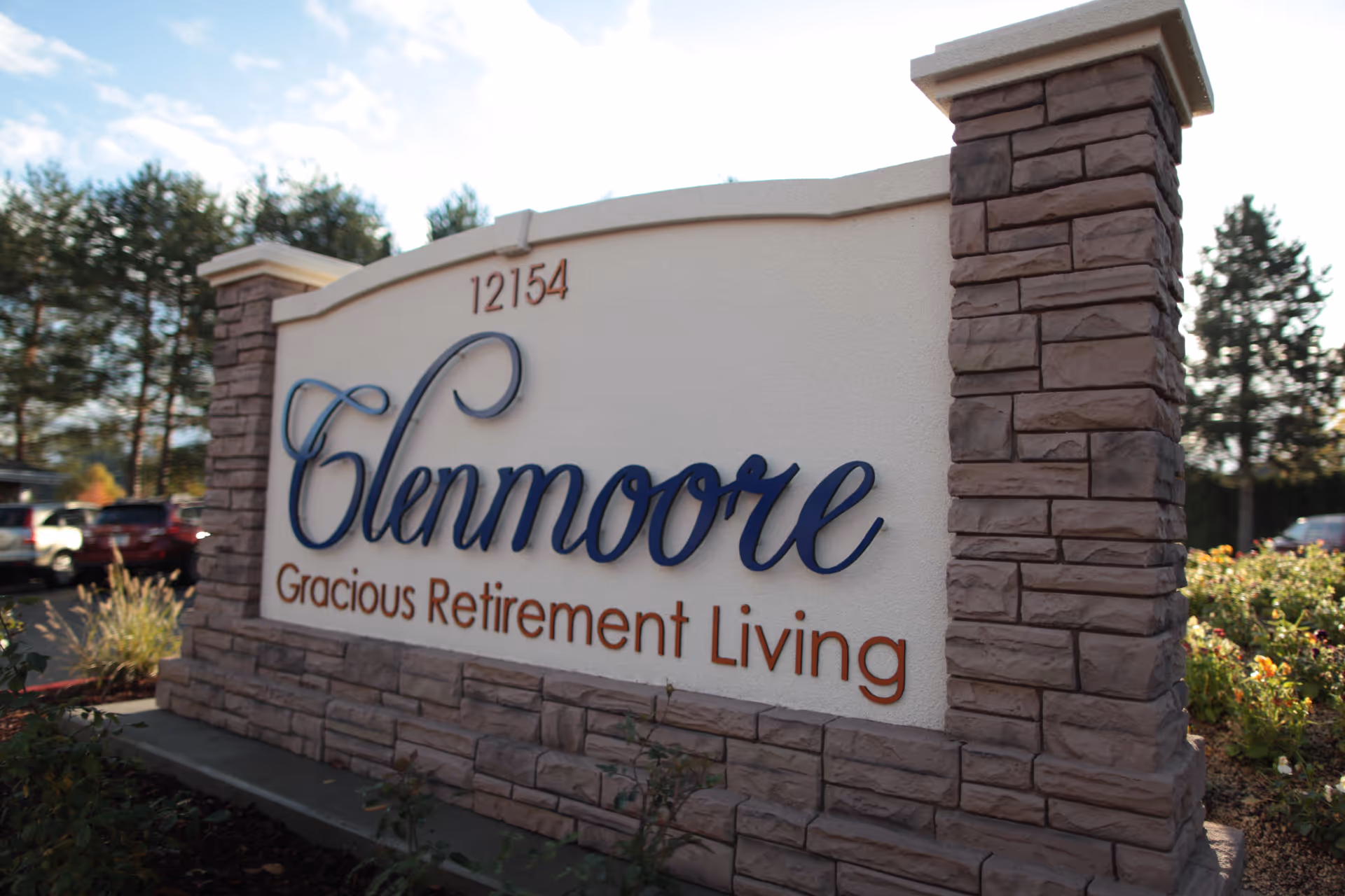Stone entrance sign reading 'Glenmoore Gracious Retirement Living' with address 12154, landscaping and parked cars behind it.