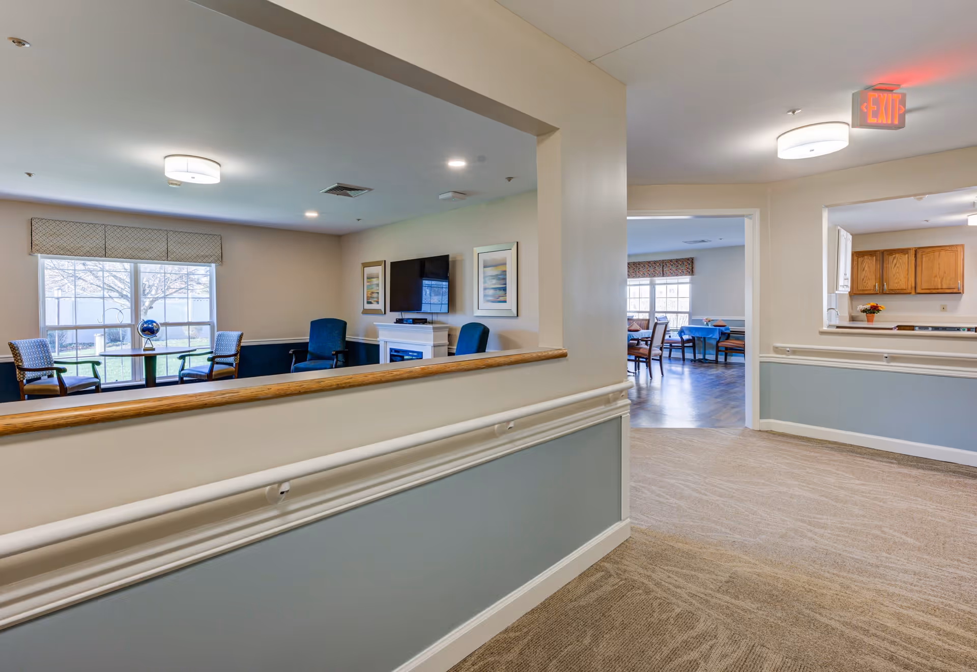 Interior view of a senior living facility hallway with beige walls and carpeted floor. To the left, there is a sitting area with chairs, a small table, a TV mounted on the wall, and framed artwork. Straight ahead, there is a dining area with tables and chairs, and to the right, a kitchen area with wooden cabinets and a pass-through window. Ceiling lights and an exit sign are visible.
