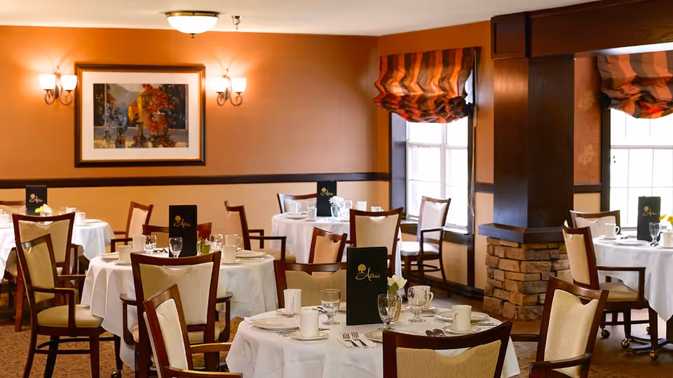 A warmly lit dining room with round tables covered in white tablecloths, set with plates, cups, glasses, and silverware. Each table has a dark menu card with the Atria logo. The room features wooden chairs with cream-colored cushions, a stone pillar, windows with striped curtains, and a framed painting on the wall.