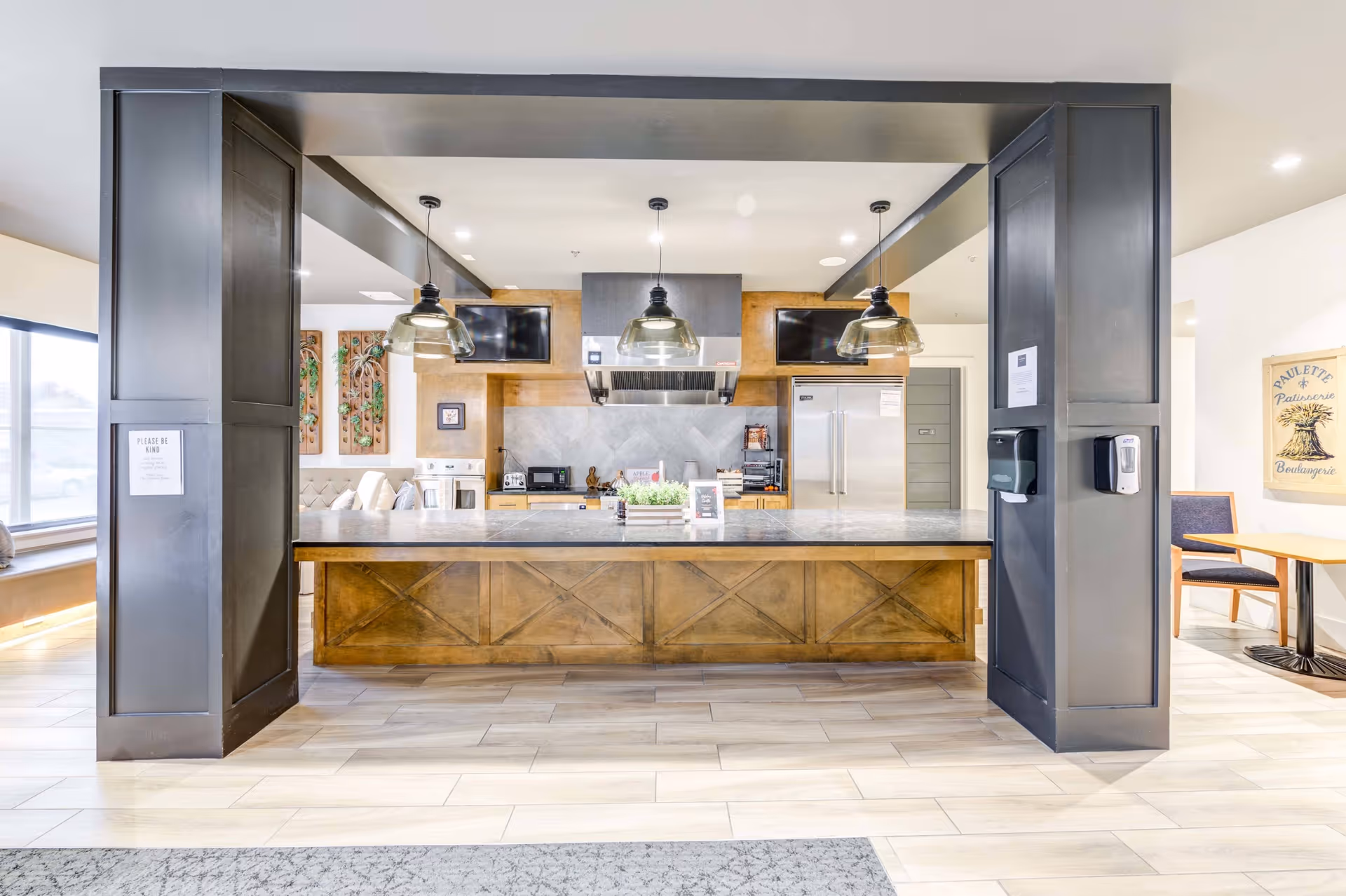 Modern kitchen area with a large island countertop made of wood and stone, three hanging pendant lights above, stainless steel appliances including a refrigerator and oven, two wall-mounted TVs, and a seating area with a table and chairs to the right.