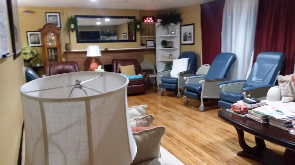 A cozy living room with wooden floors, several blue reclining chairs with wheels, a brown leather sofa, a white sofa with cushions, a wooden coffee table stacked with magazines and books, a tall grandfather clock, a large mirror on the wall, and a white bookshelf with decorative items. The room has yellow walls and red curtains covering the windows.