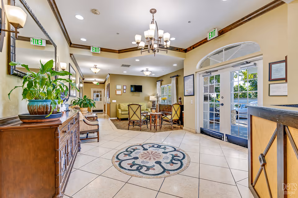 Bright and welcoming senior living facility lobby with tiled floor featuring a decorative circular mosaic in the center. There is a wooden sideboard with potted plants on the left, a seating area with chairs and a small table in the background, and glass double doors leading outside on the right. The ceiling has recessed lighting and a chandelier, with exit signs visible above doorways.
