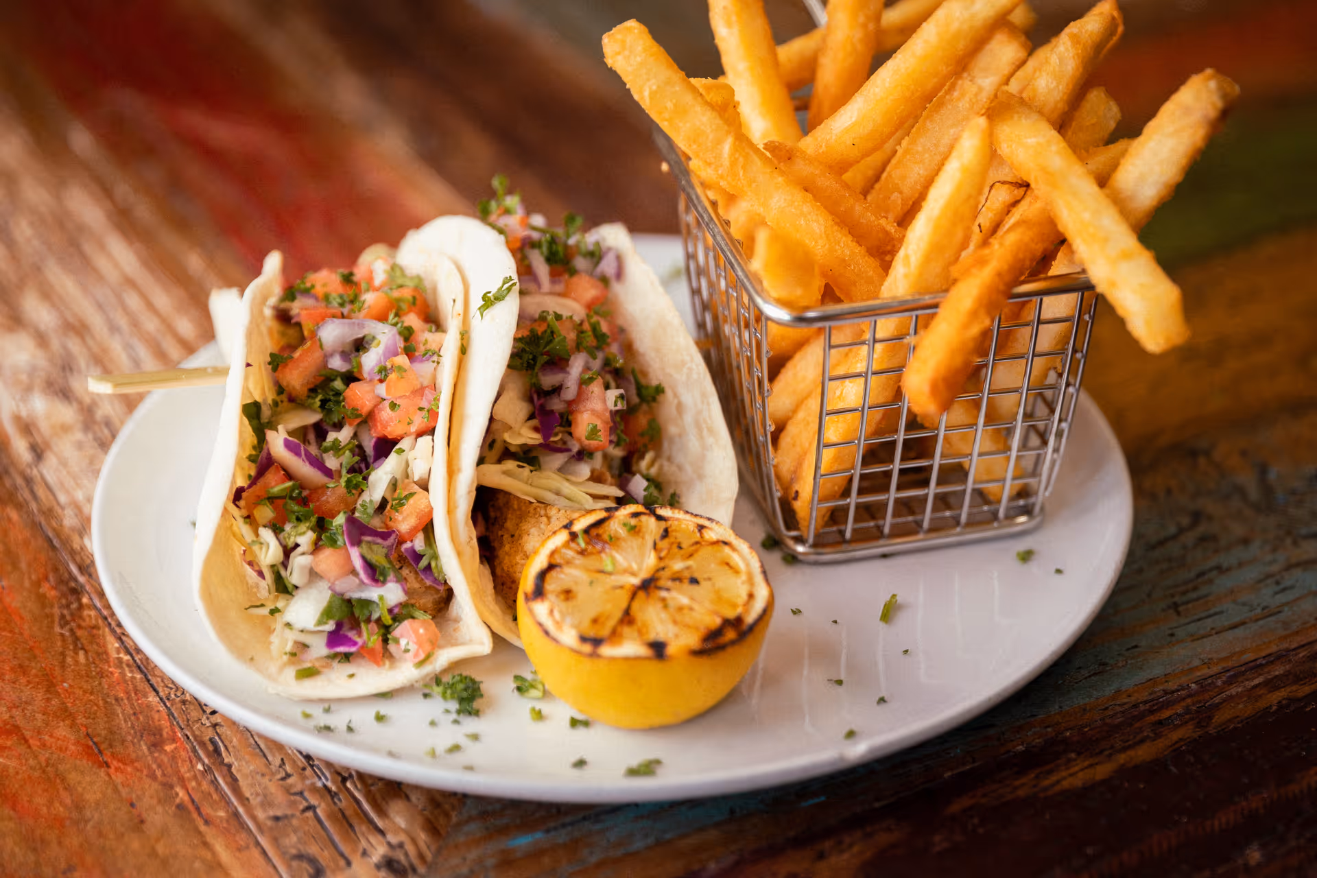 A white plate with two soft tacos filled with chopped vegetables and herbs, served with a grilled lemon half and a metal basket filled with French fries on a wooden table.