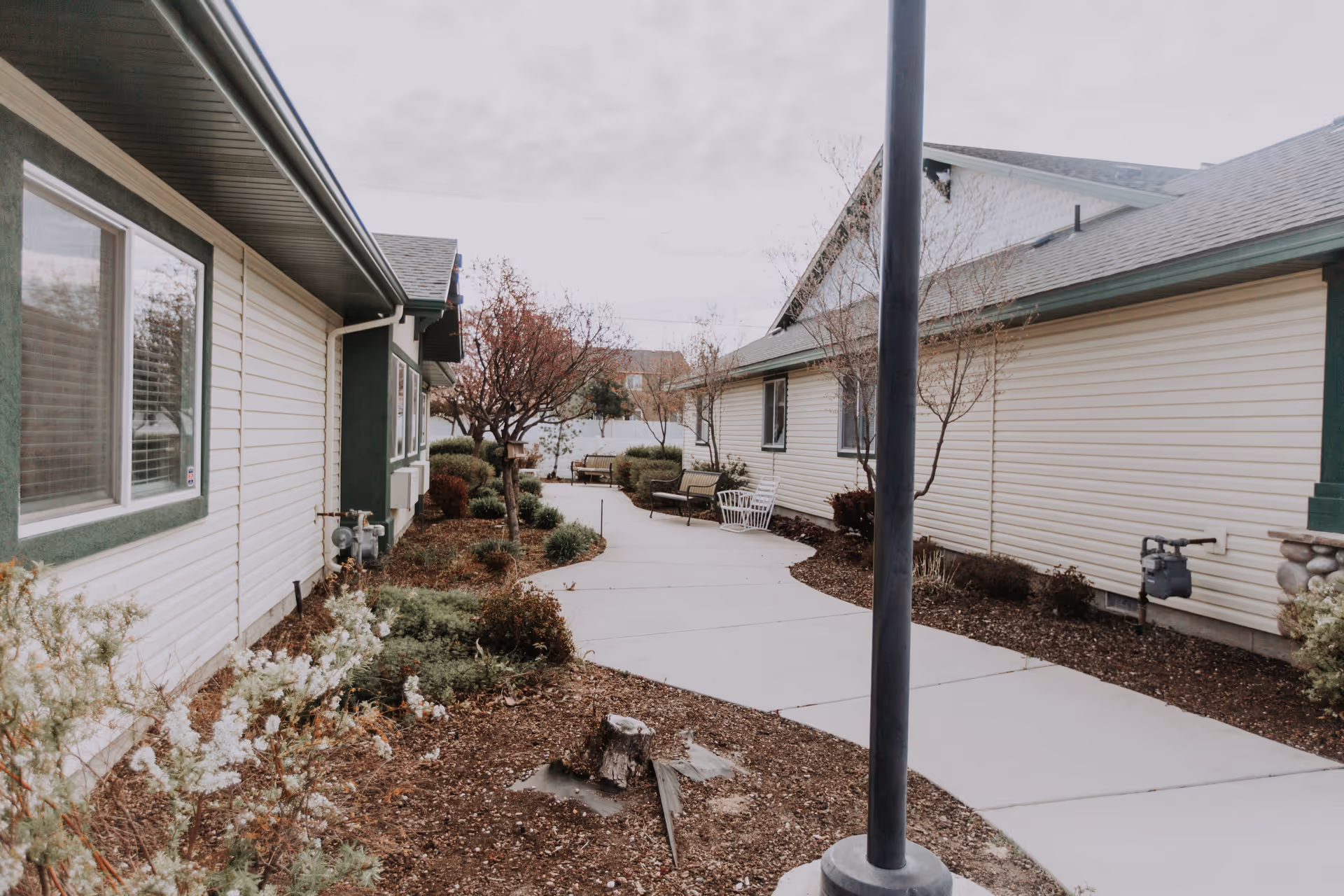Outdoor walkway between two single-story buildings with beige siding and green trim, surrounded by landscaped bushes, small trees, and benches along the path under an overcast sky.