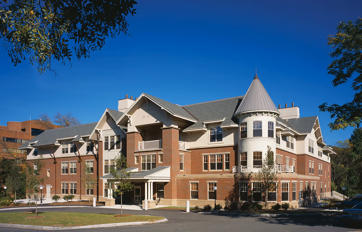 Exterior view of a large, multi-story senior living facility building with brick and light-colored siding, featuring a turret, multiple windows, and a covered entrance. The building is surrounded by trees and a clear blue sky.
