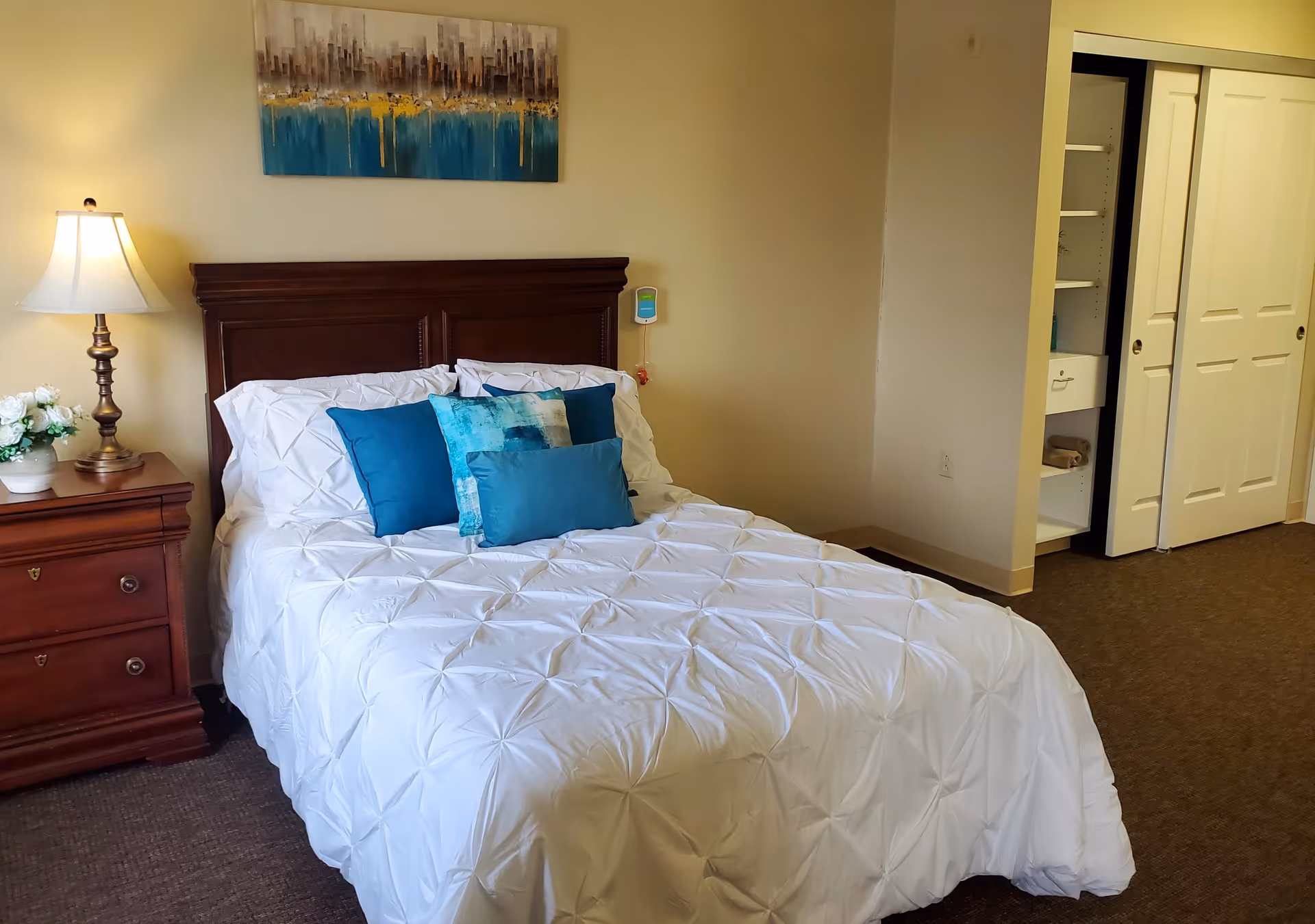 A neatly made bed with a white comforter and several blue decorative pillows in a bedroom. To the left of the bed is a wooden nightstand with a lamp and a small vase of white flowers. Above the bed hangs a colorful abstract painting. On the right side of the room, there is an open closet with white sliding doors.
