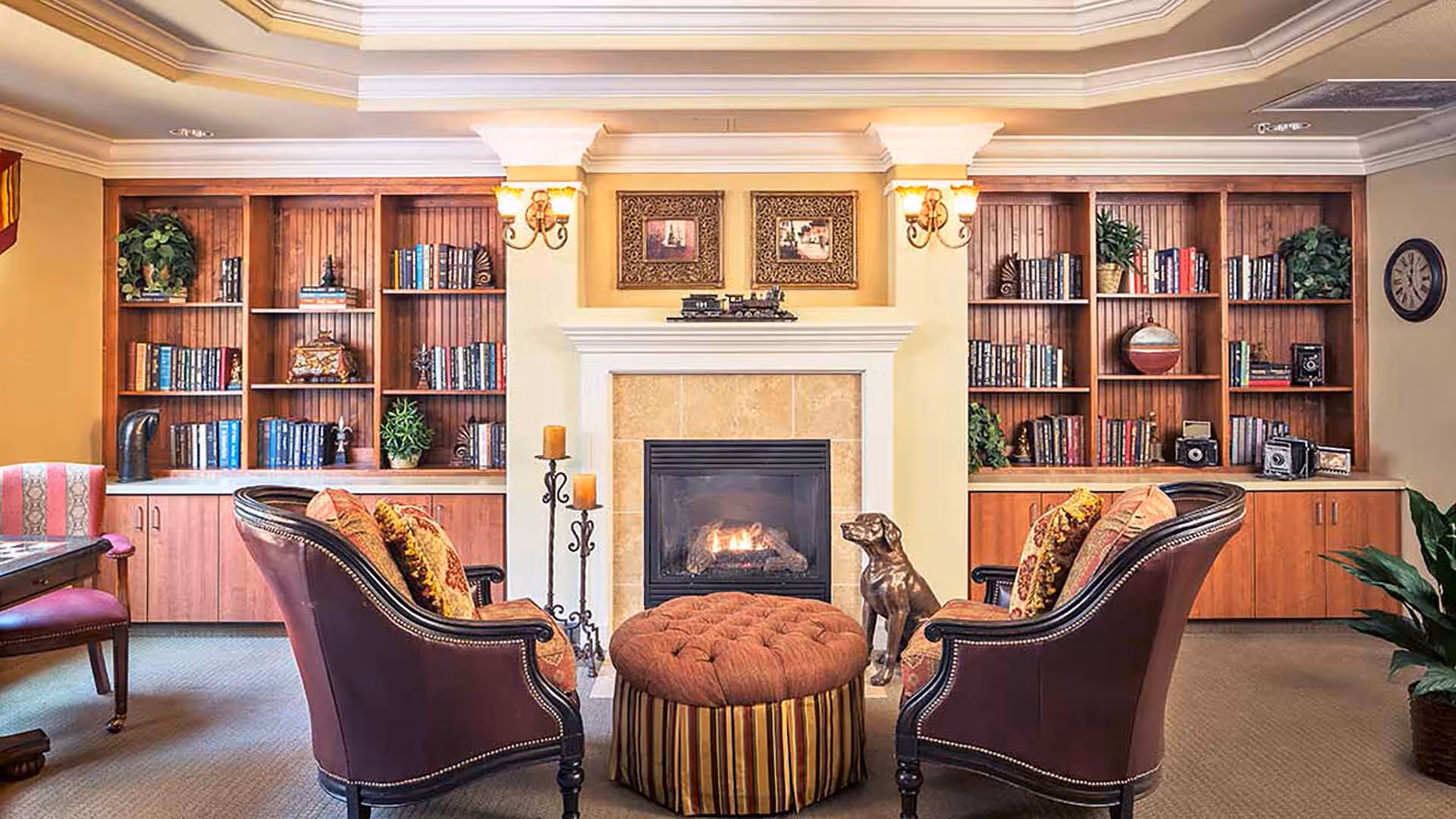 A cozy living room area with two upholstered armchairs facing a lit fireplace. Between the chairs is a round, tufted ottoman with striped fabric. Behind the fireplace are built-in wooden bookshelves filled with books, plants, and decorative items. The room has warm lighting with wall sconces and a clock on the right wall.