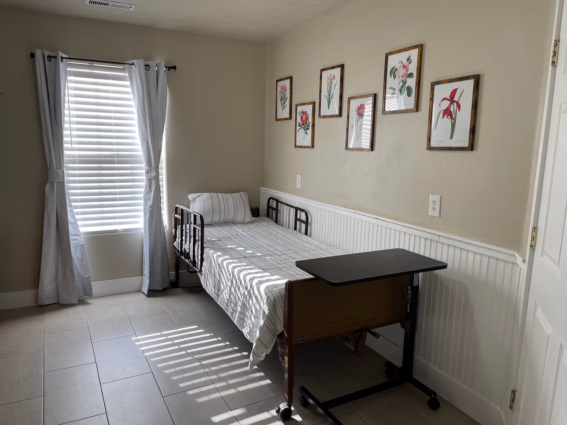 A simple, clean bedroom with a single bed featuring a metal frame and striped bedding. Next to the bed is an adjustable overbed table on wheels. The room has beige walls with white wainscoting and six framed botanical prints hanging above the bed. A window with white blinds and light gray curtains allows sunlight to stream into the room, casting shadows on the tiled floor.
