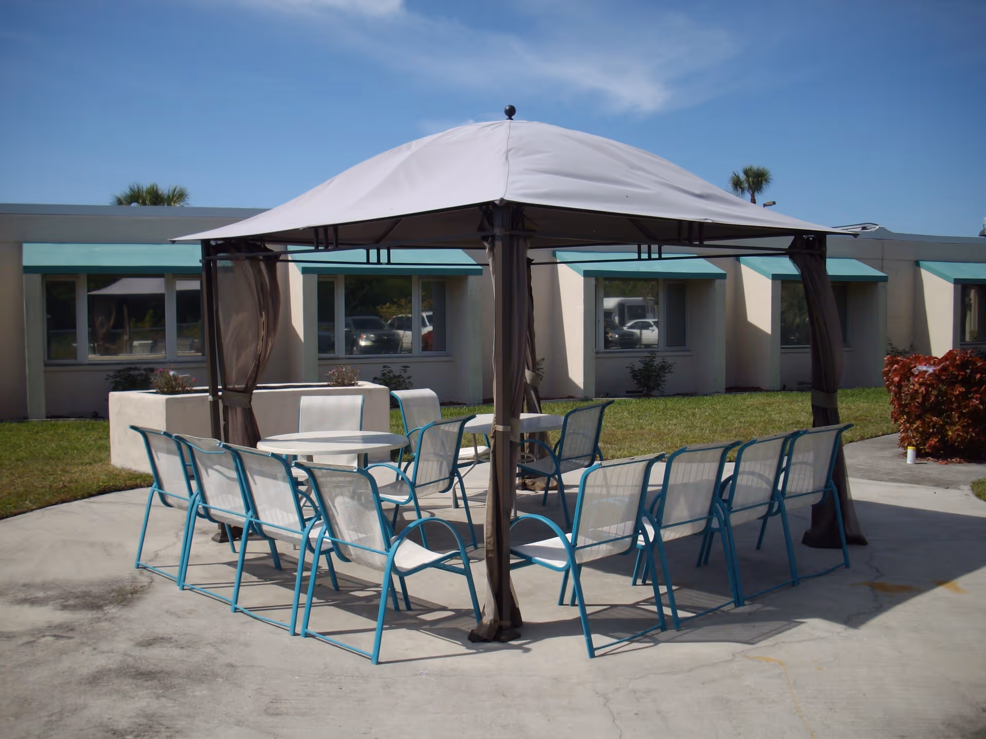 Outdoor seating area with a canopy covering a round table surrounded by multiple blue and white chairs on a concrete patio. The background shows a single-story building with windows and green awnings, along with some grass and shrubs under a clear blue sky.